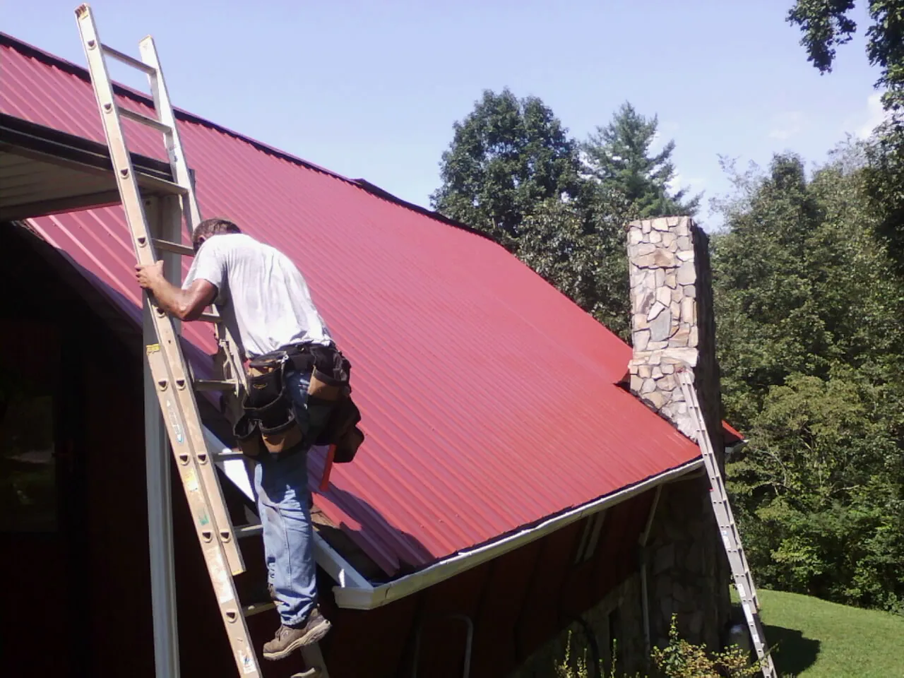 Roofer on a ladder beside a red metal roof and stone chimney — Soffit & Fascia Repair by Roofing Brothers in Bighorn